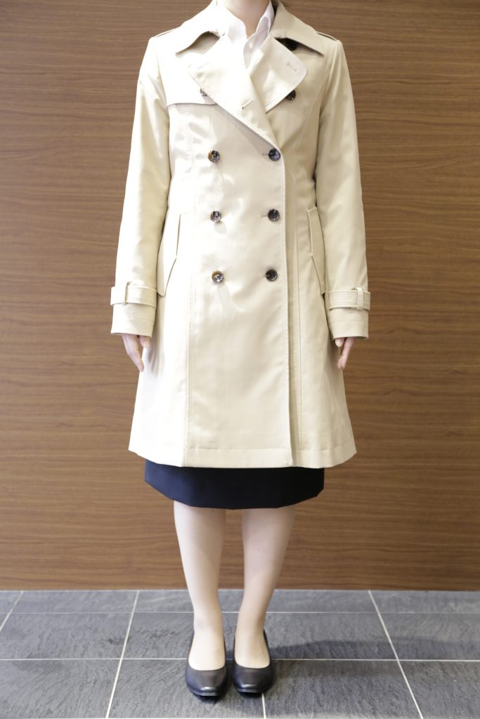 ベージュトレントコートを着用した女性が、こちらを向いて立っている画像。