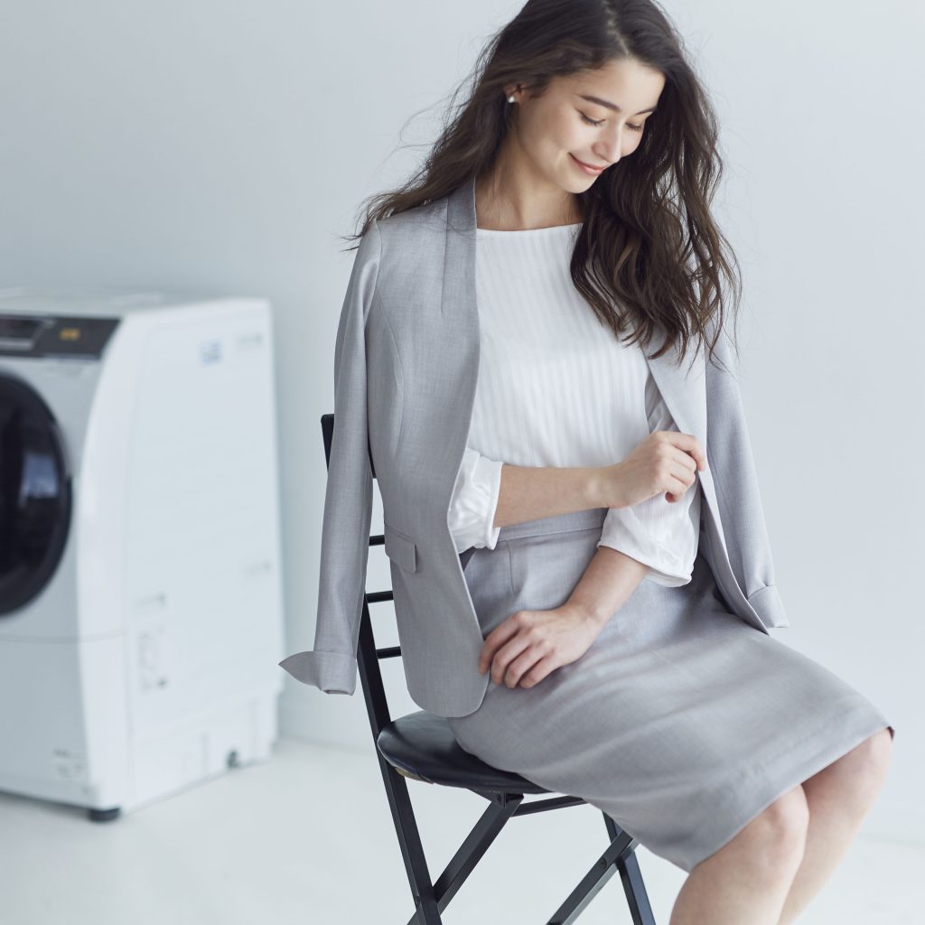 洗濯機の前でライトグレーのノーカラーのスカートスーツを着用した女性が座っている。
