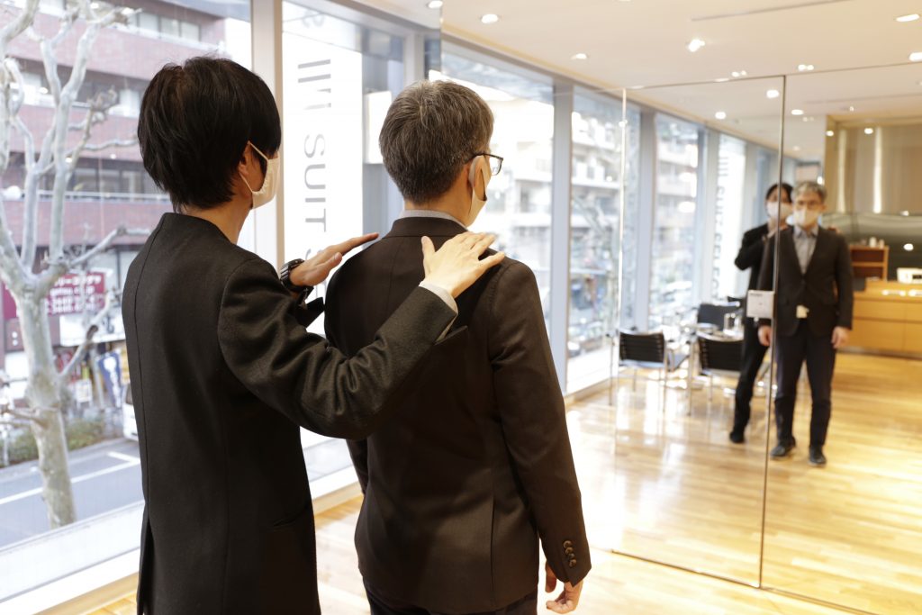 スタッフが、全身鏡を利用しながら、お客様にオーダースーツの接客をしている画像。
スタッフがお客様の背後から、両肩に手を添えている。