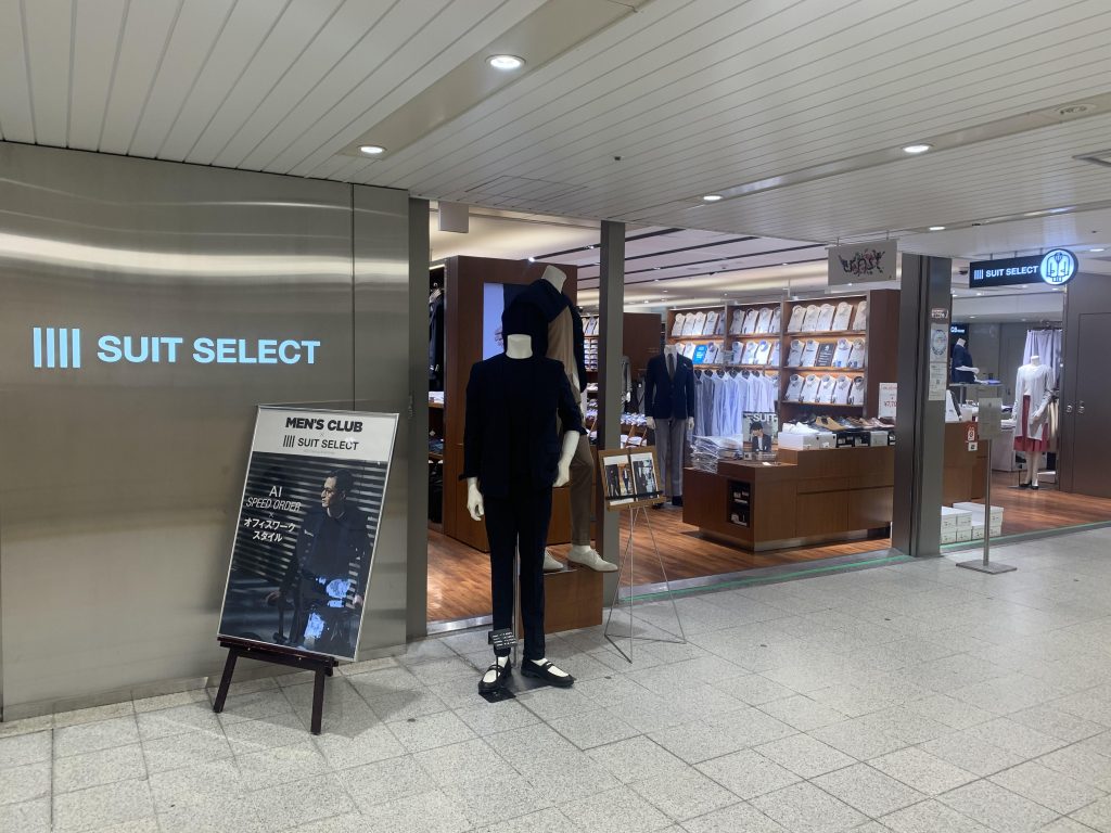 スーツセレクト大阪駅クロスト店の店頭画像。中央、ブラックスーツ着用のマネキンが立っている。右手奥に、シャツの陳列棚。