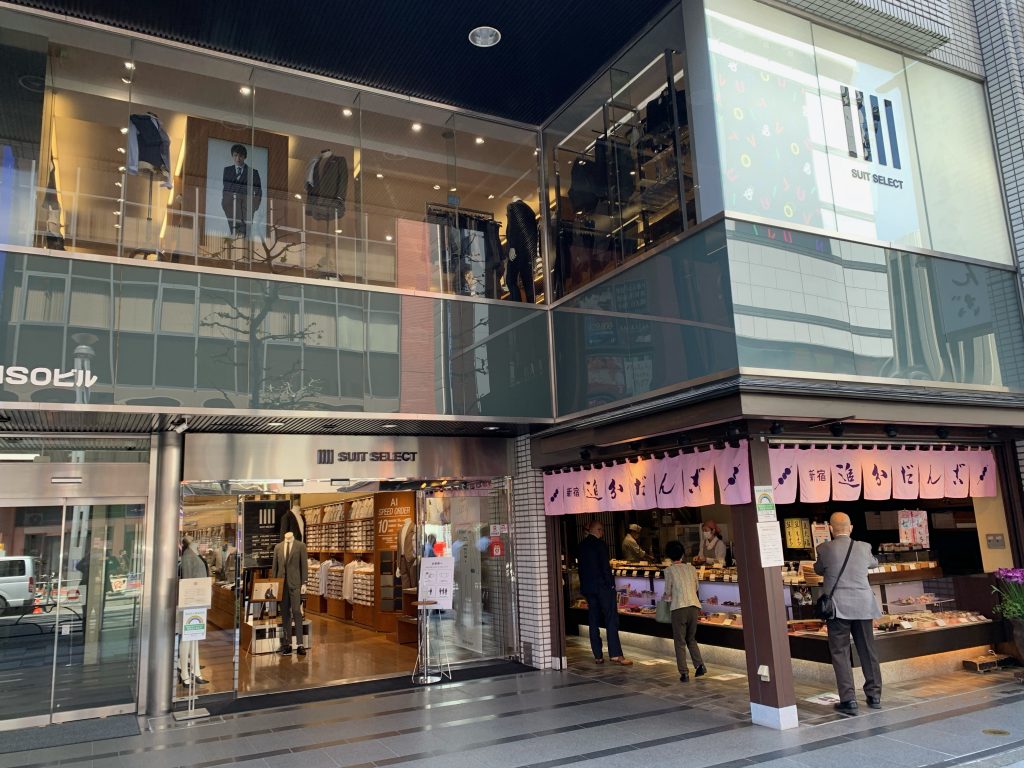 スーツセレクト新宿店の店頭画像。右手は追分団子やさん。ガラス張り。入口頭上に、SUIT　SELECT　のロゴが設置されている。