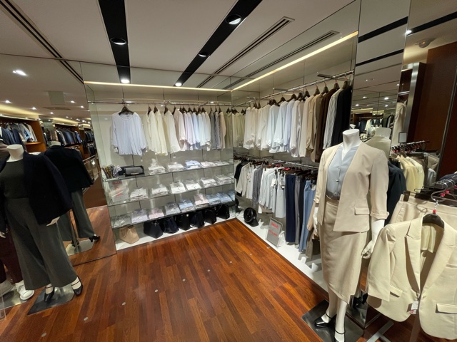 新宿南店　店内画像。　レディースコーナー。ベージュのスカートスタイルスーツを着用したマネキンが右手に。左側にはグレー系のパンツスタイルのマネキンが立っている。中央にはブラウスやインナーの陳列棚。