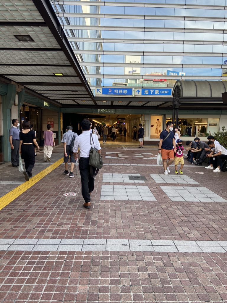 横浜駅西口バスロータリー画像。数人が行き来。駅ビルの下の通路。煉瓦調のタイル。ガラス張りの駅ビル。