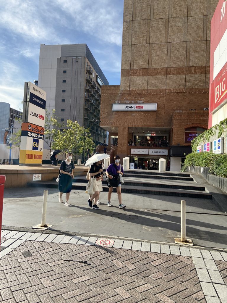 スーツセレクト横浜西店店頭広場の画像。奥のほうに横浜西店の入り口が見える。手前はグレーの階段になっていて、ちょっとしたスペースになっている。4人の女性グループがその広場を横切っている。