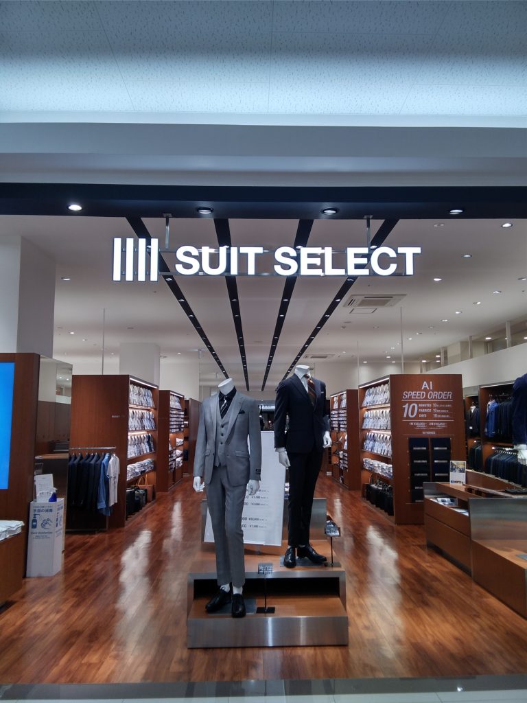 ゆめタウン光の森の店頭の様子。
ダーク系のスーツと、グレースーツを着た2体のマネキンが中央に立っている。その奥は、店内の商品が整然と陳列されている。マネキンの頭上には、suit　selectのロゴ。