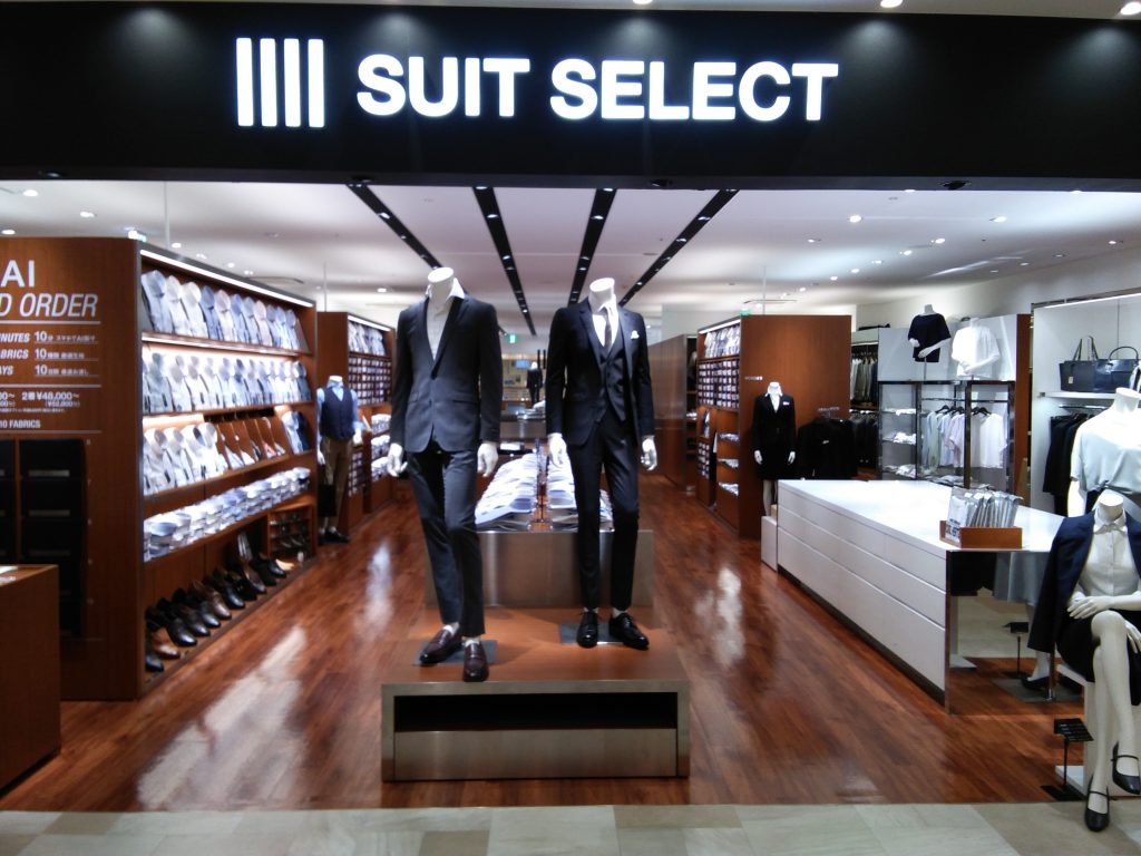 サクラマチ熊本の店頭の画像。
中央にダーク系のスーツを着た2体のマネキンが立っている。右側はレディースコーナー。左側には豊富なシャツの陳列。