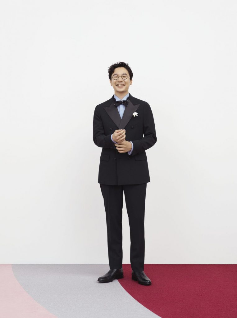 前原さんが、左手で右手のカフスを正すようなしぐさで、こちらを見て微笑んでいる。全身の画像。
服装は、ブラックのダブルのスーツスタイル。淡いブルー系のシャツ、ニット素材のボウタイを合わせている。胸元には白い花。
背景はほぼ白。足元だけ、ピンク、グレー、ワインの床。