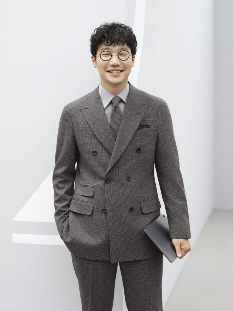 前原さんがチャコールグレーのダブルのスーツを着用して、こちらを見て微笑んでいる。同じくグレー系のシャツとネクタイを合わせている。
右手はパンツのポケットの中、左手にはipadのようなモバイルを持っている。膝から上の画像。白い壁面の部屋の一角のような場所。