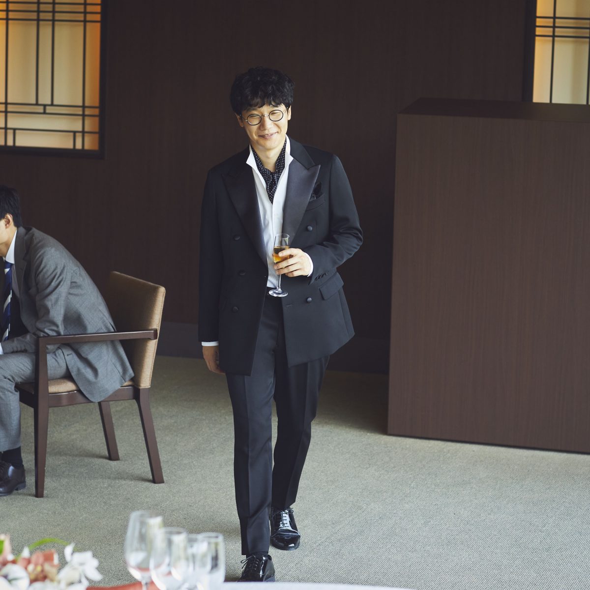 前原さんが左手にシャンパングラスをもって、微笑んでこちらに歩いてきているような全身の画像。服装はブラックのダブルのスーツ。ホワイトシャツの首元のボタンを外して、ネイビー系のスカーフデザインのタイを合わせている。ブラックシューズ。何かのパーティー会場でのワンシーンのような画像。
背後に、ブラウンチェアに腰かけているグレースーツスタイル男性1名が確認できる。