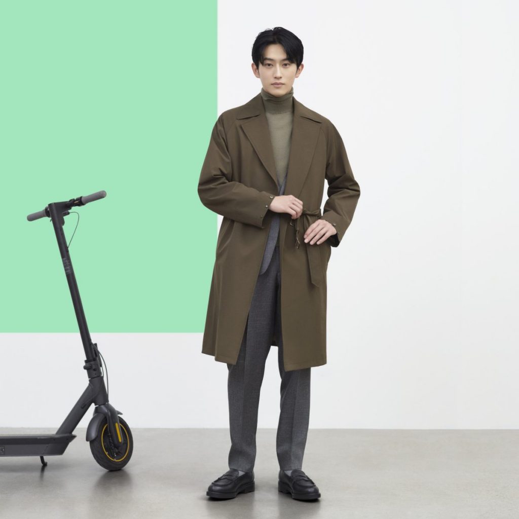 杉野さんがブラウン系タートルネックニット、グレー系パンツ、JK,ブラウン系コートを着用して、こちらをみて立っている画像。靴は黒。黒色の電動キックボードが杉野さんの後方左奥に見えます。