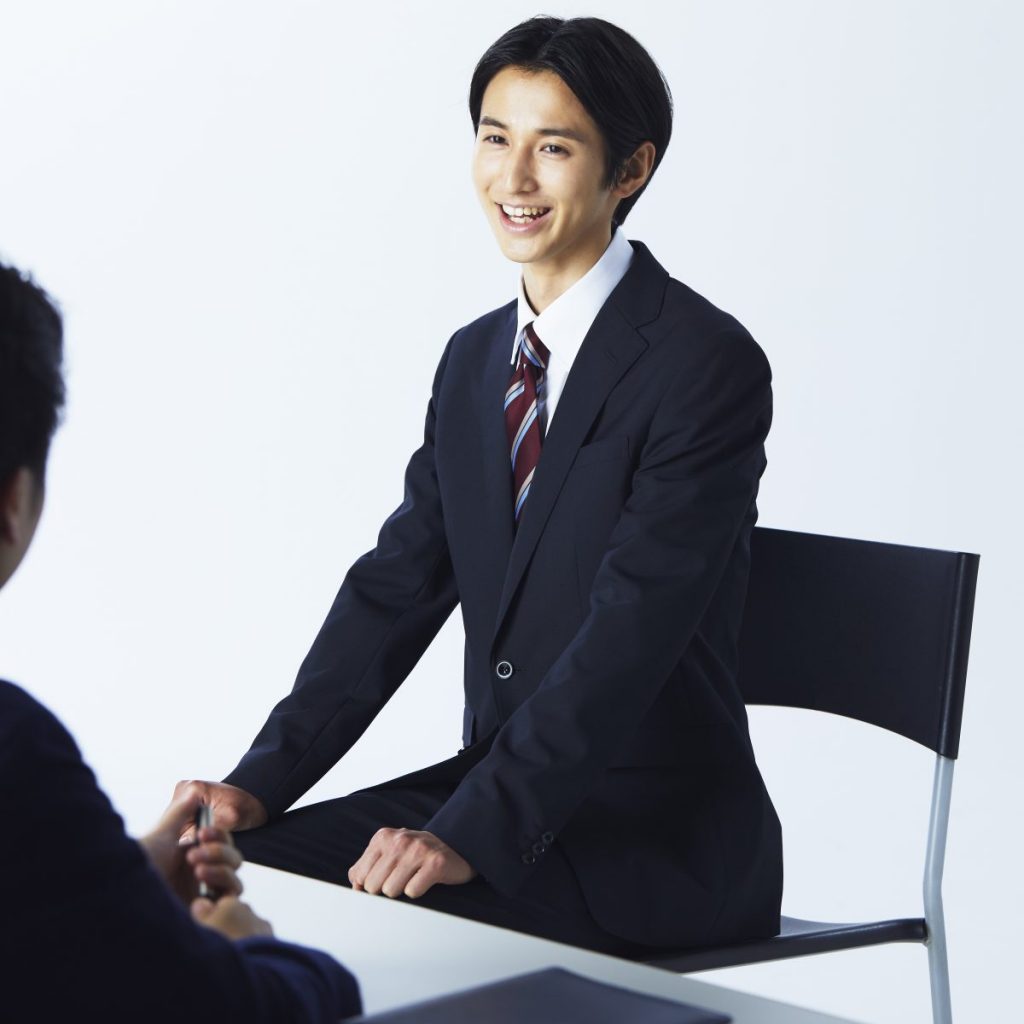 リクルートスーツスタイルの男性が、椅子に腰かけて面談中の様子。エンジ系ストライプネクタイ着用。